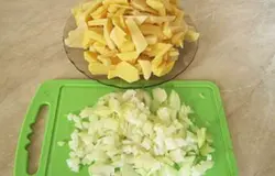 Тщательно щеточкой помыть картофель. С картофеля срезать кожуру, выложить на разделочную доску и нарезать тоненькими колечками. Очистить от шелухи репчатый лук, ополоснуть водой и нарезать полукольцами. Разморозить готовое тесто, распаковать и выложить на стол, присыпанный мукой. Разделить тесто примерно на две одинаковые части. Раскатать в тонкие пласты.