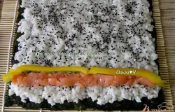 На специальный бамбуковый коврик положить лист нори, сверху выложить часть риса, распределить мокрыми руками по всему листу (оставив 2-3 см с одного края). Посыпать рис кунжутом, выложить полоски лосося и нарезанный длинной соломкой перец.